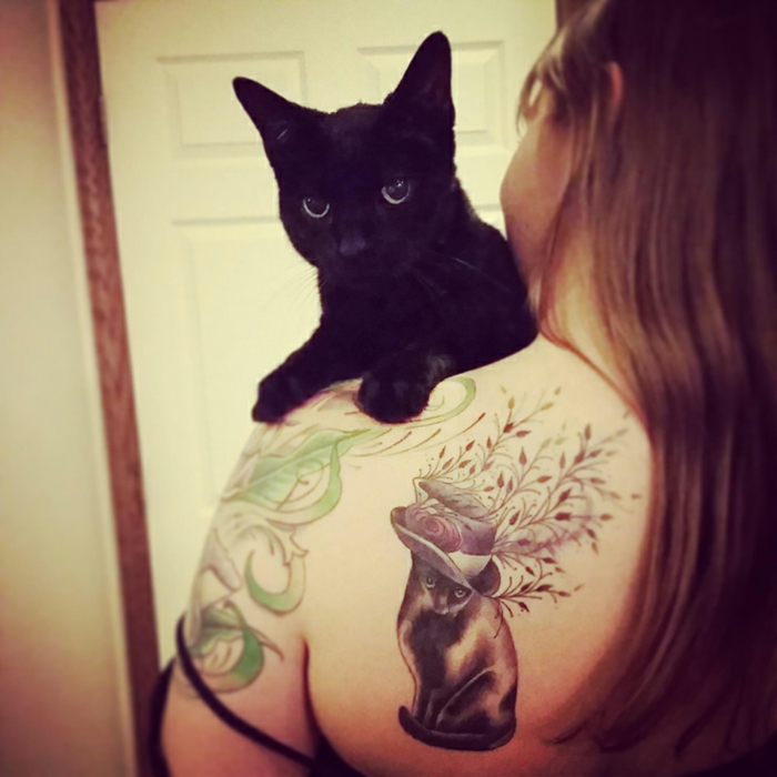 Täältä löytyy ajatus kissan tatuoinnille - tässä on musta kissa ja nainen, jonka tatuointi olkapäällä on musta kissa, hattu ja kasvit