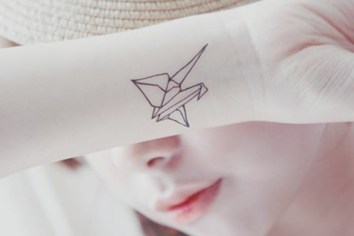 Tässä on nuori nainen, jolla on pieni origami-tatuointi rannalla - pieni lentävä origami-lintu kädessä