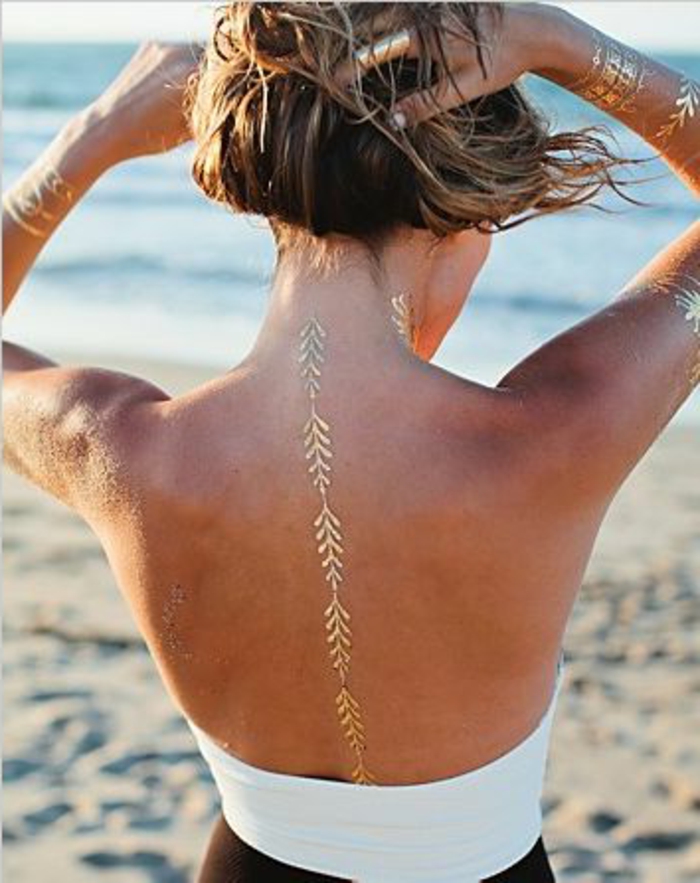 татуировка идеи фина линия в златист цвят на гърба на жена с руса коса на плажа