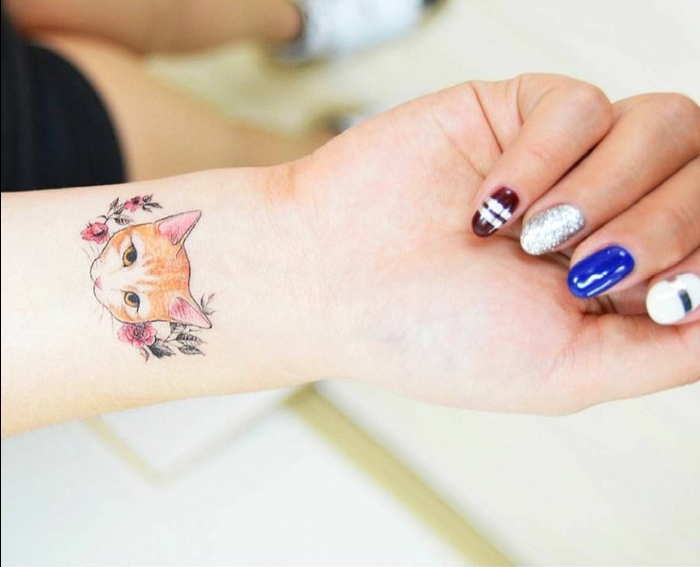 A kéz, az ujj, a színes körömlakk, a macska tetoválás a csuklóján, egy kis macska, vörös virágok